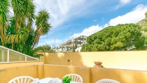 Una mesa lista para el desayuno, con sillas blancas, una pared amarilla y vistas a una colina con edificios.