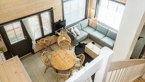 Une pièce aux hauts plafonds, dotée d’un plancher en bois, d’une table ronde en bois entourée de chaises, d’un canapé gris et d’une grande fenêtre avec des stores.