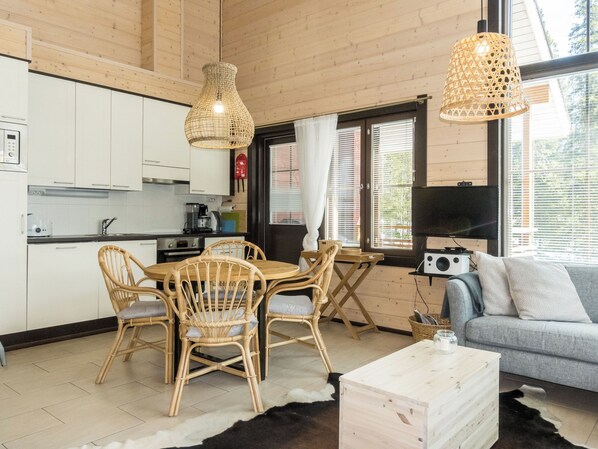 A modern kitchen with white cabinets, a dining area with wicker chairs, a grey sofa, and a television.
