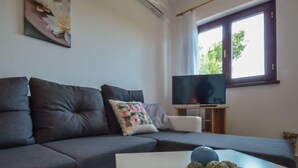 Una sala moderna con un sofá gris oscuro, una mesa de centro blanca con objetos decorativos, un televisor de pantalla plana sobre un soporte de madera y una ventana con cortinas.