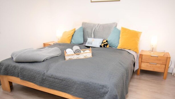 A neatly made bed with a grey quilt, a wooden nightstand with a lamp, and a bedside table with a basket of folded towels. - Vacation home Butzel by Interhome (Norddeich)
