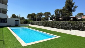 Une piscine rectangulaire bordée de blanc, entourée de gazon synthétique vert, le tout se détachant sur un ciel bleu clair.
