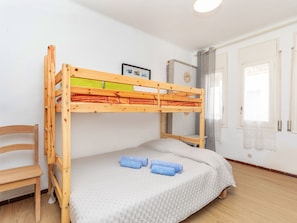 A bunk bed with a wooden frame, a single bed below, a chair, a framed picture on the wall, and a window with curtains.