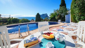 Ein Poolbereich mit Meerblick, ein Tisch mit Essen und einem Buch sowie helle Liegestühle.