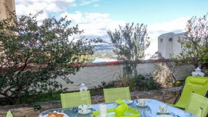 A glass table with a tray of food, surrounded by green chairs, set against a scenic backdrop with a view of a body of water and a white building. - Hameau la Madrague by Interhome (Saint Cyr sur Mer La Madrague)