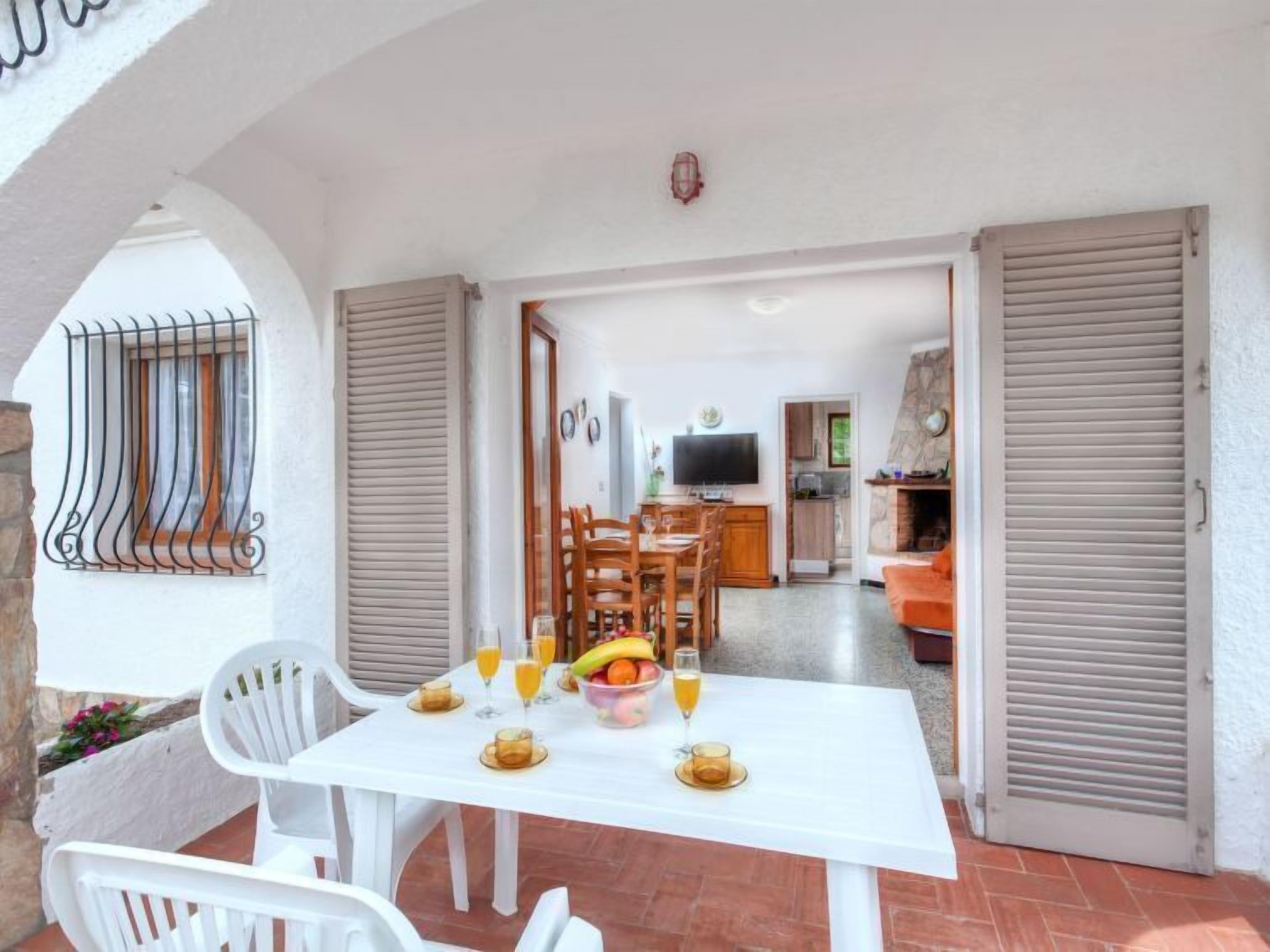 A dining area with a table set for four, a kitchen in the background, and a balcony with white chairs.
