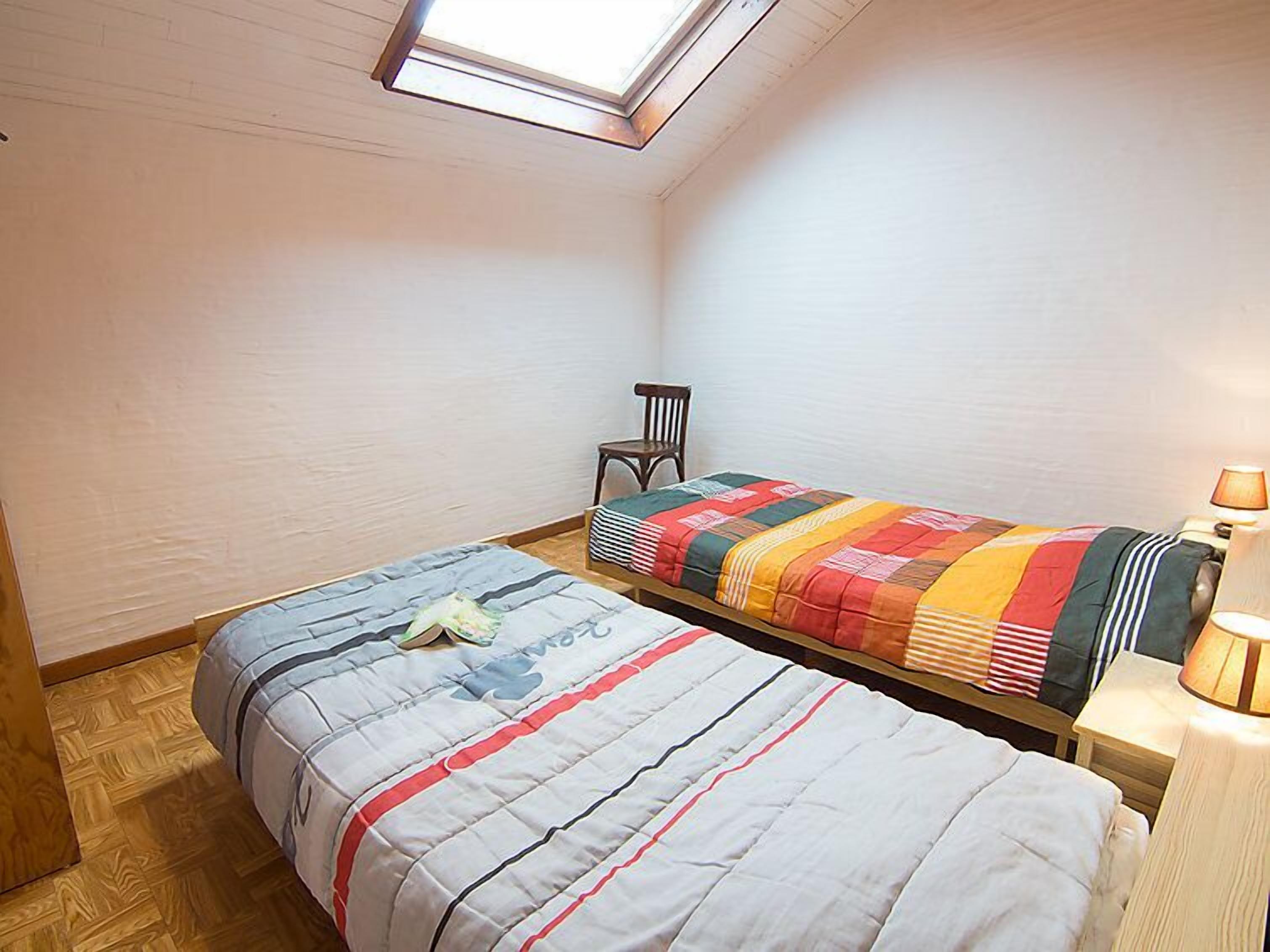 Un dormitorio pequeño y acogedor con dos camas, un tragaluz, una silla y una mesita de noche con lámpara.