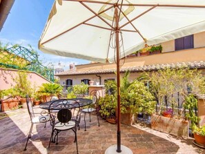 Une terrasse extérieure avec une table ronde et des chaises, entourée de plantes en pot et d’un parasol blanc.
