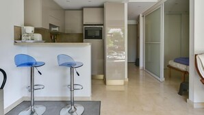 Uma cozinha moderna com uma ilha central, dois banquetes azuis e vista para uma área de jantar com divisória de vidro.