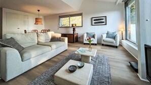 Ein modernes Wohnzimmer mit einer Couch, zwei Sesseln, einem Couchtisch und einem Beistelltisch. Ein Fenster mit Blick auf Bäume und eine Tür im Hintergrund sind zu sehen.
