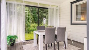 Une terrasse couverte avec une table et des chaises, de grandes fenêtres avec des rideaux légers, une plante en pot et une vue sur la verdure.