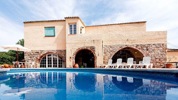 Un espace aménagé au bord de la piscine, avec un bâtiment en pierre, des portes en arc de cercle et des sièges d’extérieur.