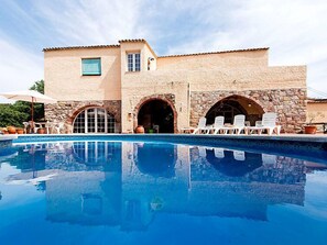 Un espace aménagé au bord de la piscine, avec un bâtiment en pierre, des portes en arc de cercle et des sièges d’extérieur.