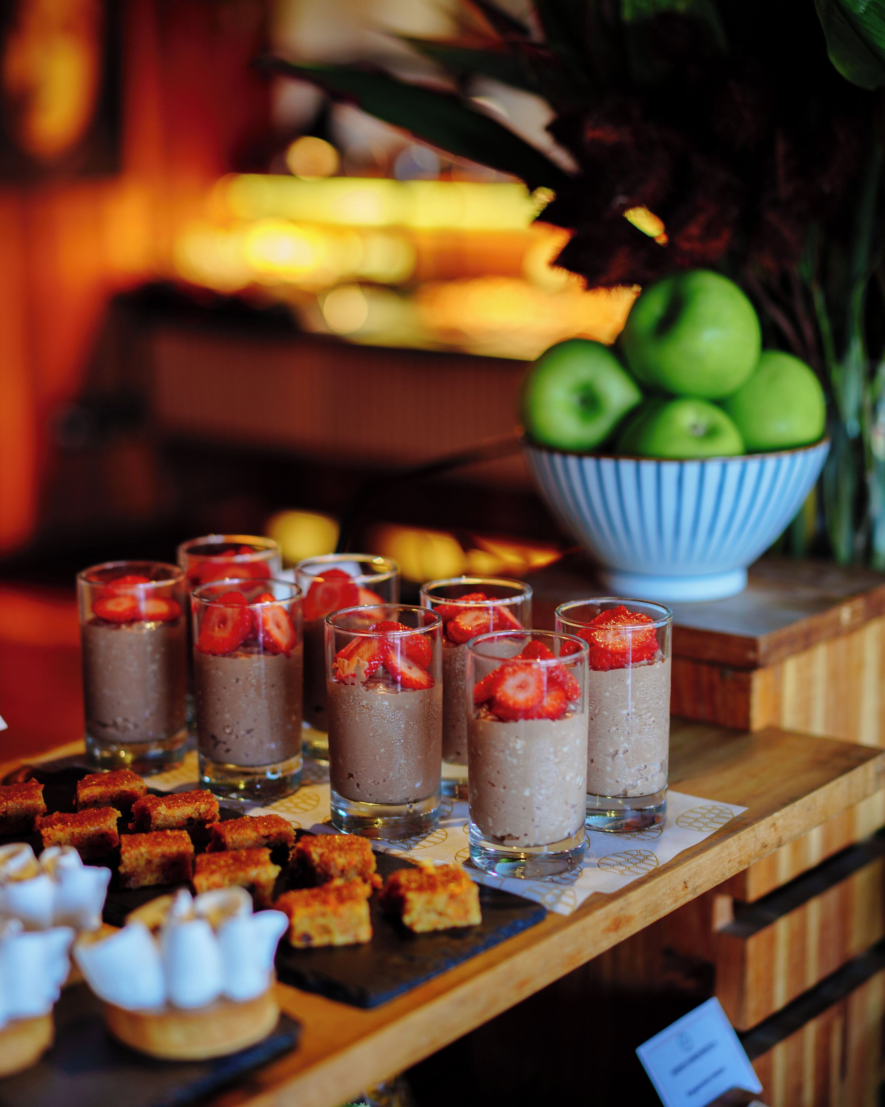 Una mesa de postres con vasitos de mousse de chocolate cubiertos de fresas, un bol de manzanas verdes y pequeños pasteles.