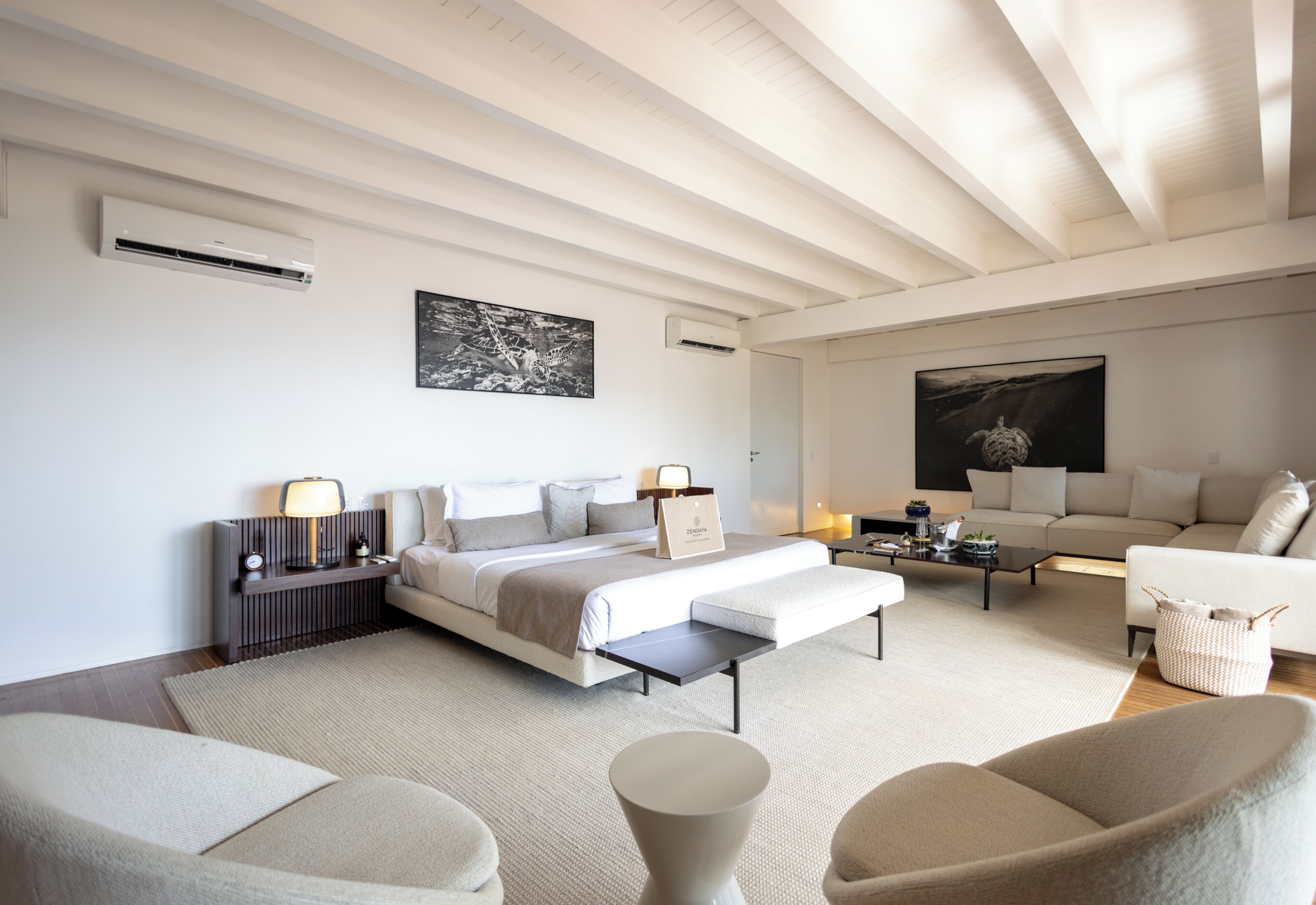 A modern hotel room with a large bed, a sofa, a coffee table, and a chair. There are air conditioning units on the walls and a painting above the bed.