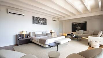 A modern hotel room with a large bed, a sofa, a coffee table, and a chair. There are air conditioning units on the walls and a painting above the bed.