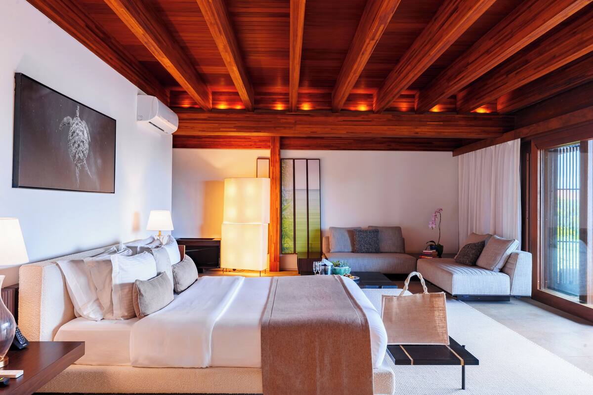 Une chambre d’hôtel moderne avec un plafond en bois, un grand lit, un canapé, une table basse et une vue sur l’extérieur.