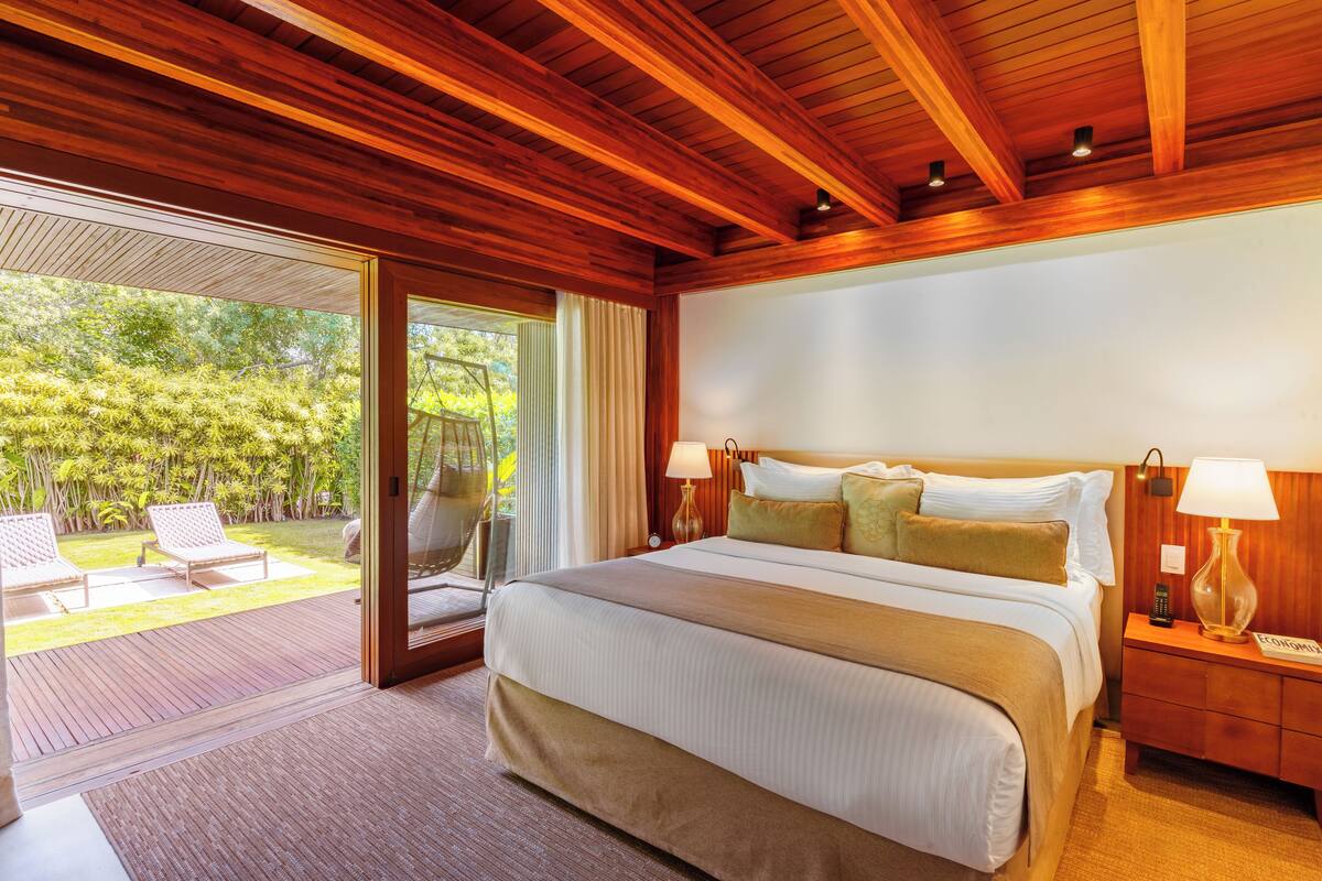 Une chambre spacieuse avec un grand lit, des poutres apparentes en bois au plafond, une table de chevet avec une lampe et une vue sur un jardin luxuriant, visible à travers une porte-fenêtre coulissante.