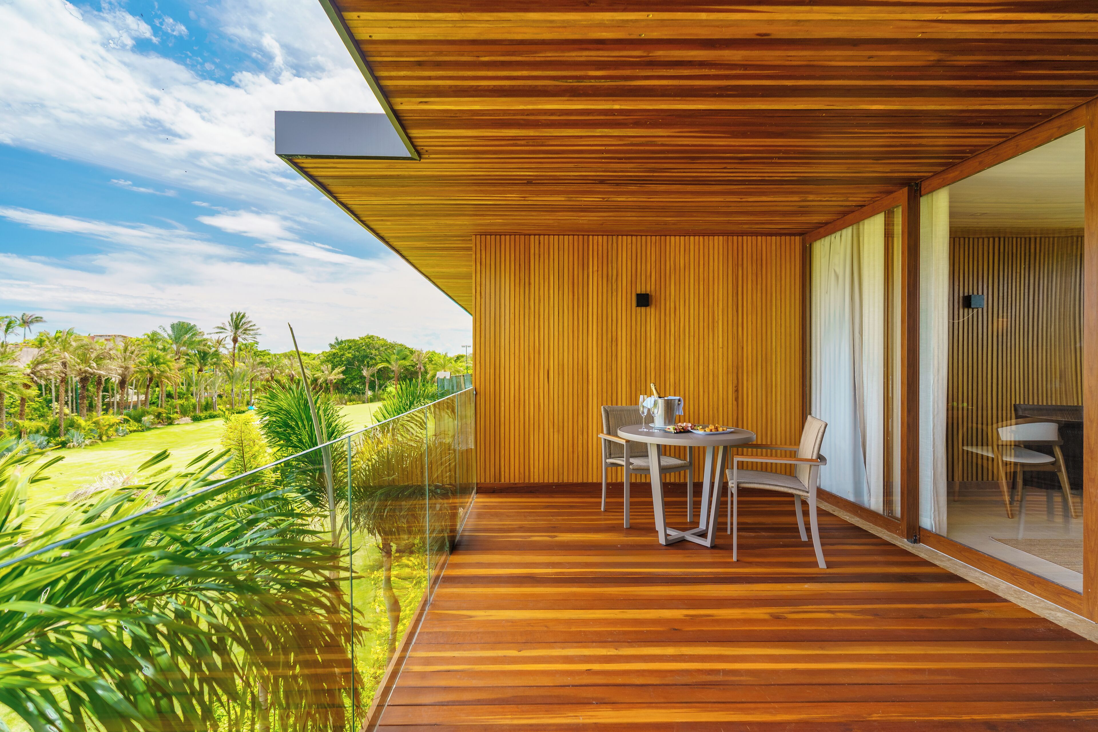 Um deck de madeira com mesa e cadeiras, com vista para um jardim exuberante e um céu límpido.