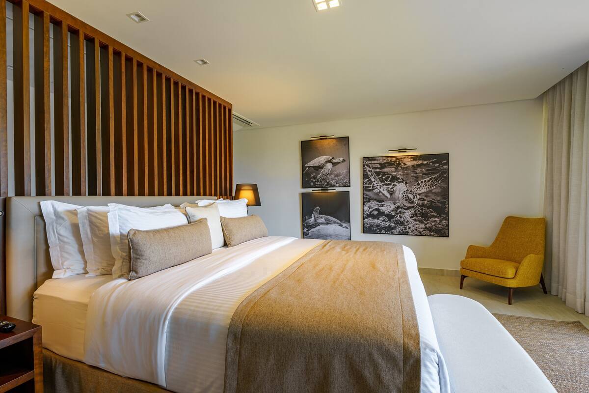 Une chambre d’hôtel avec un grand lit, une tête de lit en lattes de bois, un fauteuil jaune et deux tableaux encadrés au mur.