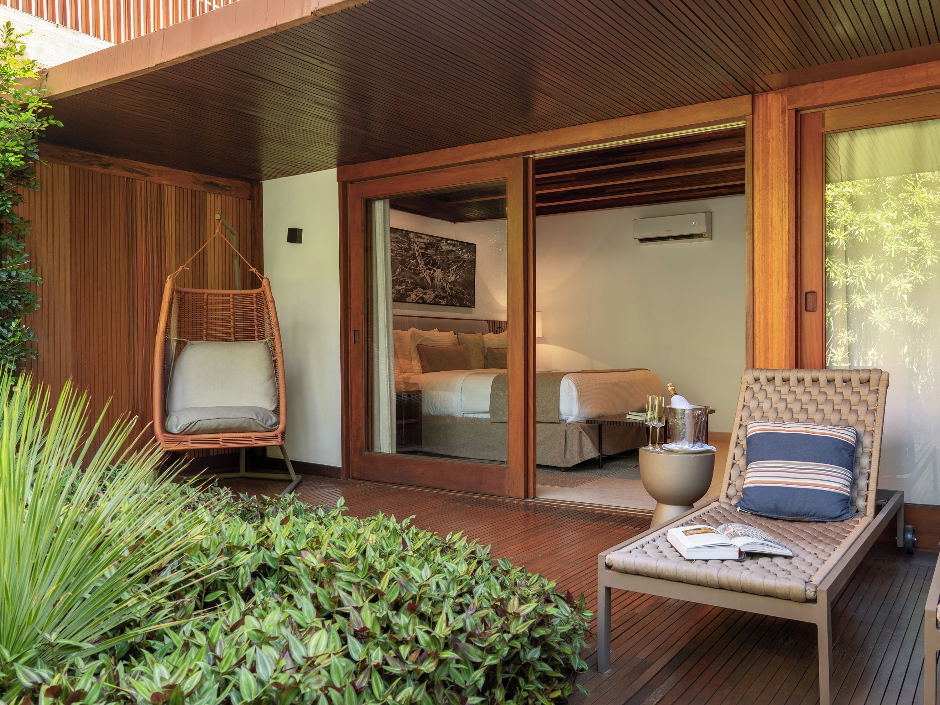 A wooden deck with a hanging chair, a lounge chair with a book, and a view into a bedroom with a bed and a nightstand.