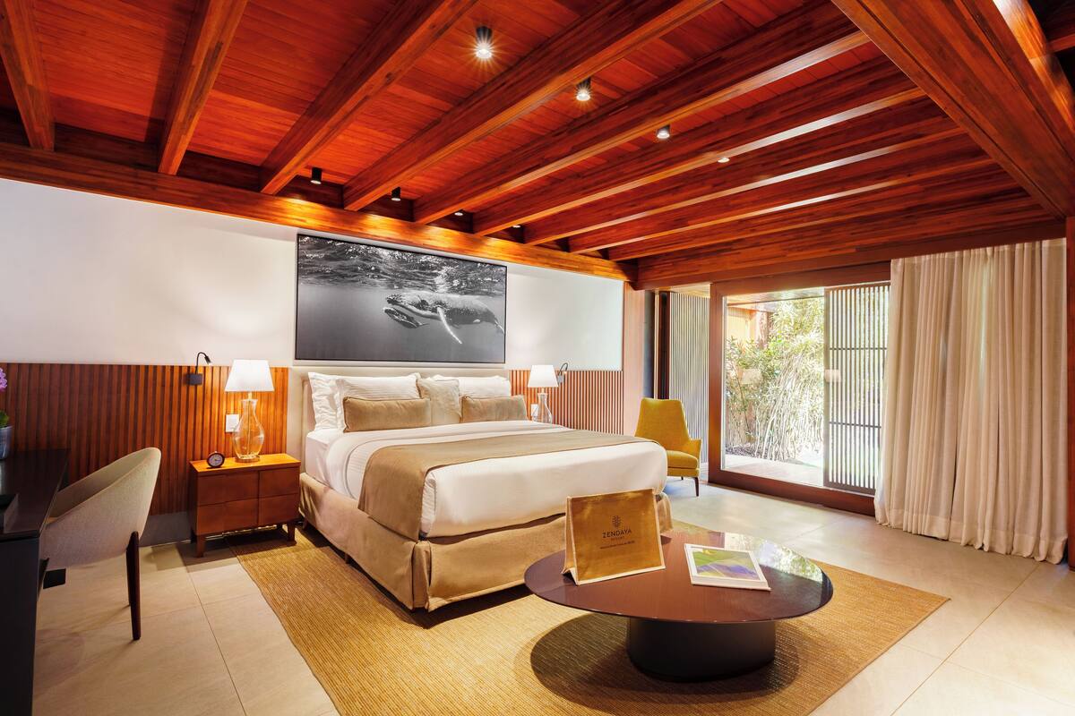 Une chambre d’hôtel avec un grand lit, des poutres apparentes en bois au plafond, un bureau avec une chaise, une table de chevet, une lampe, une table basse et une grande fenêtre avec des rideaux.