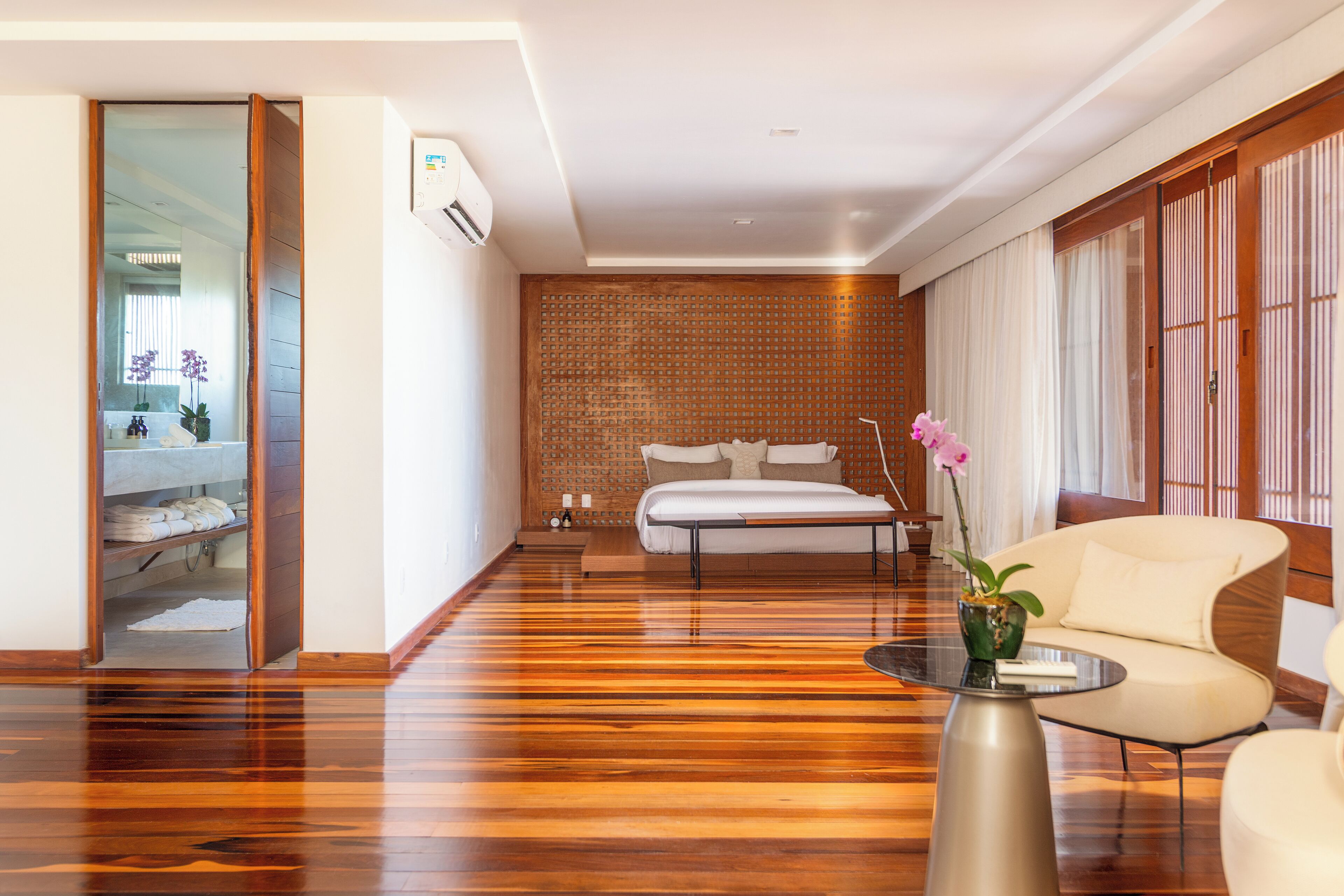 A modern hotel room with a wooden floor, a sofa, a small table with a vase, and a sliding door leading to a bathroom.