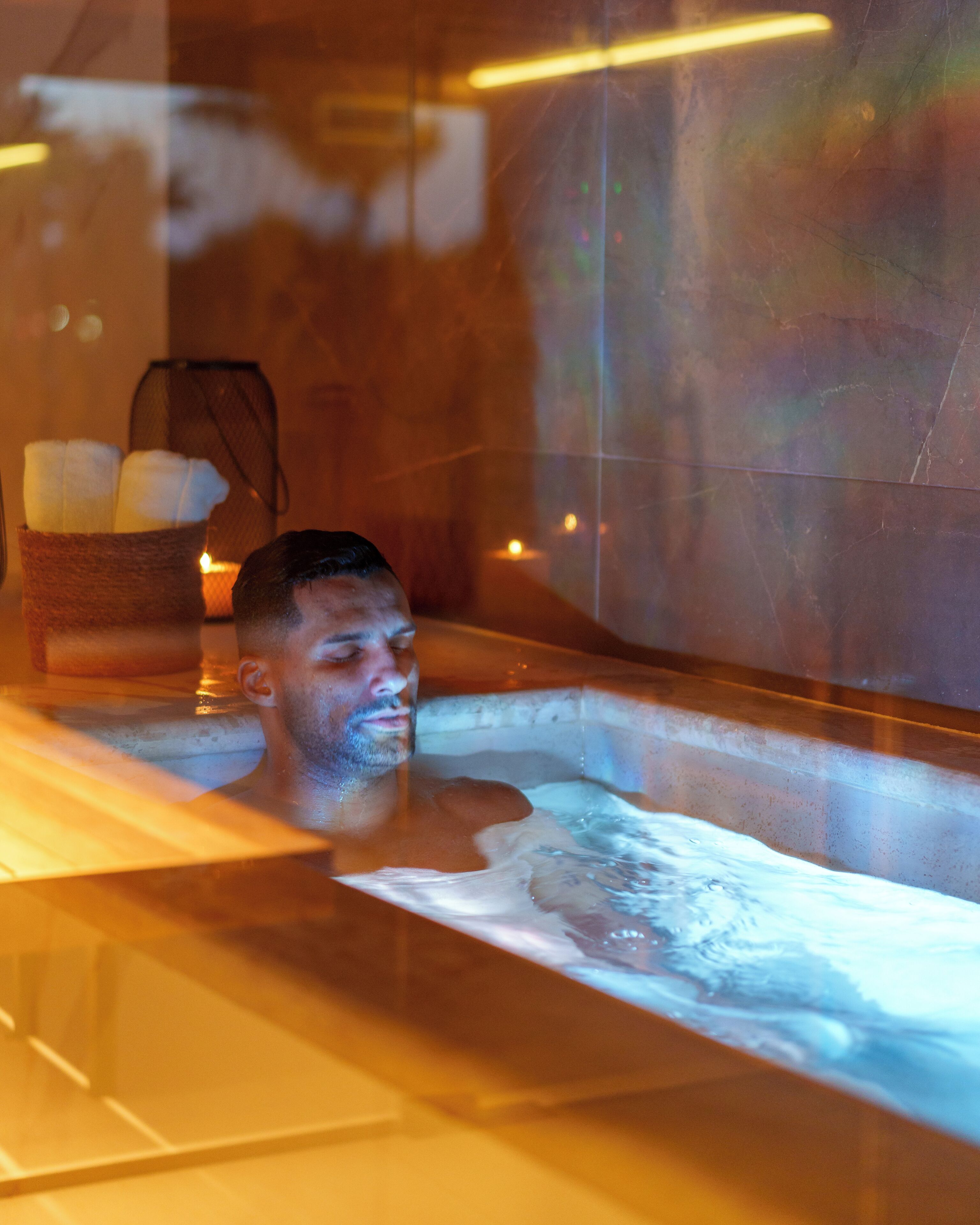 Une personne dans un bain à remous, avec une bougie et un panier rempli de serviettes.