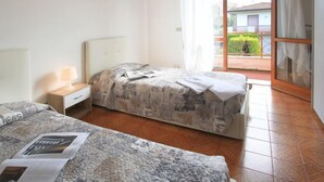 A bedroom with two beds, a nightstand with a lamp, a window with curtains, and a view of a garden. - Marcella by Interhome (Forte dei Marmi)