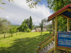 A wooden play structure with a red roof and a blue signboard in a grassy area with trees and a distant view of houses.
