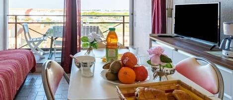 A balcony with a bed, a table with fruits, a basket of bread, a TV, and a window with curtains.