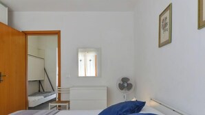 Un dormitorio con cama, mesita de noche, silla, una pequeña mesa, ventana, ventilador y puerta.