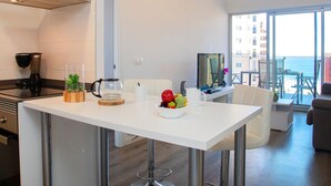 A modern kitchen with a white countertop, a coffee maker, a kettle, and a bowl of fruit. There is a dining area with a white table and chairs, and a TV on a stand. The room has large windows with a view of the ocean. - Mirador de Calpe by Interhome (Calpe/Calp)