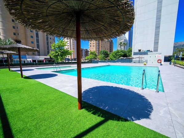 A swimming pool with a thatched umbrella, surrounded by artificial grass and a cityscape in the background.