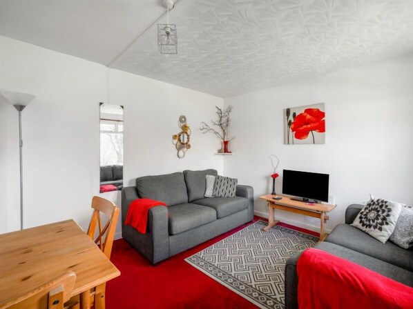 A modern living room with a grey sofa, a wooden dining table, and a TV on a wooden stand.