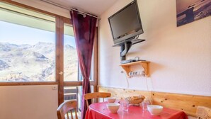 Ein Essbereich mit rotem Tischläufer, Holzkästen, einem an der Wand befestigten Fernseher und Blick auf eine gebirgige Landschaft durch ein Fenster.