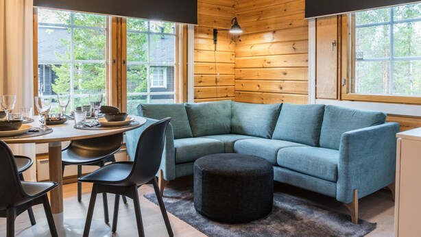 A cozy cabin interior with a wooden ceiling, a teal sofa, a dining table with chairs, and large windows offering a view of greenery outside.