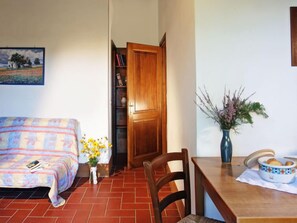 A cozy living room with a sofa, a dining table with chairs, a wooden door, a bookshelf, and a vase with flowers.