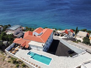 A coastal hotel with a swimming pool, red-roofed buildings, and a view of the sea.