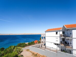 A coastal building with balconies, a clear view of the sea, and a hilly landscape in the background.