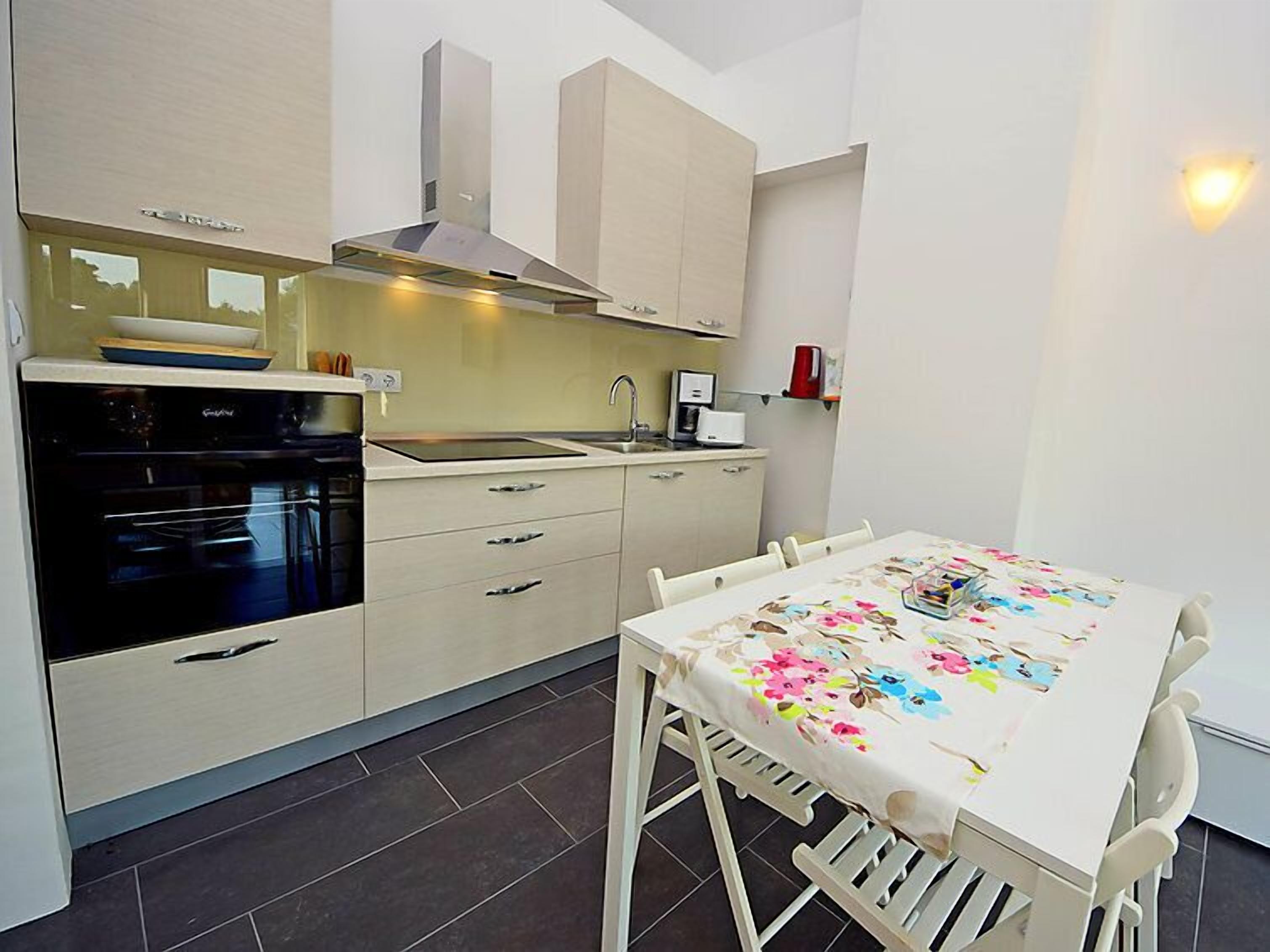 A modern kitchen with a built-in oven, a stove, and a sink. There is a dining table with a floral tablecloth and chairs.