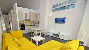 Un salon moderne avec un canapé jaune vif, une petite table blanche et un coin cuisine à l’arrière-plan.