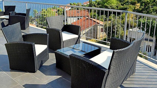 Un balcon doté de meubles en osier et d’une table en verre, offrant une vue sur la côte et les montagnes en arrière-plan.