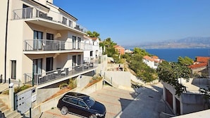 Un immeuble résidentiel à plusieurs étages, doté de balcons, avec une voiture garée devant, et offrant une vue sur la mer et les montagnes en arrière-plan.