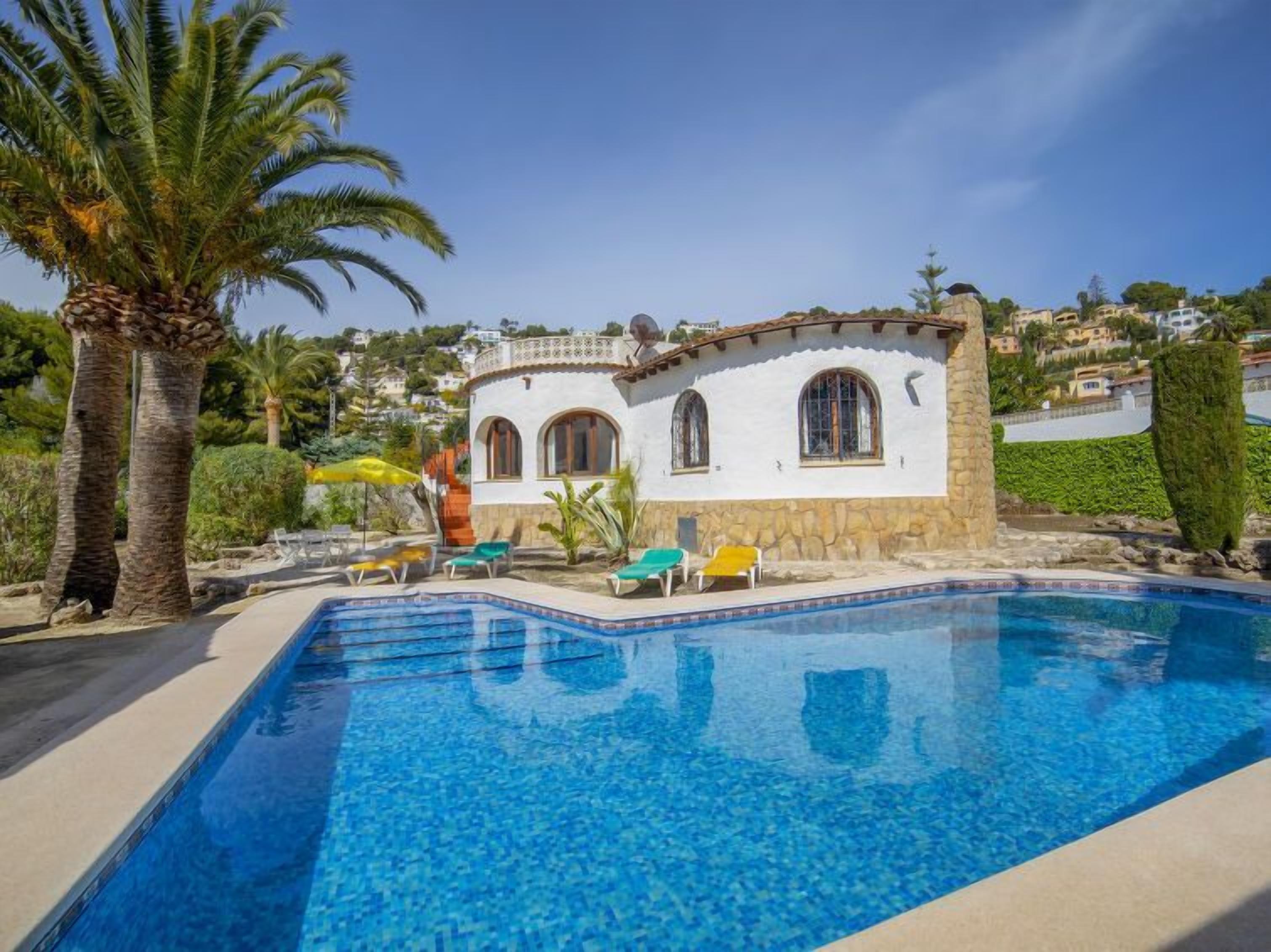 A poolside view of a Mediterranean-style villa with a clear blue pool, palm trees, and a stone terrace.