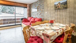 Un coin repas avec une table en bois dressée pour quatre personnes, des coussins rouges sur un canapé et un mur en pierre orné d’un tableau encadré.