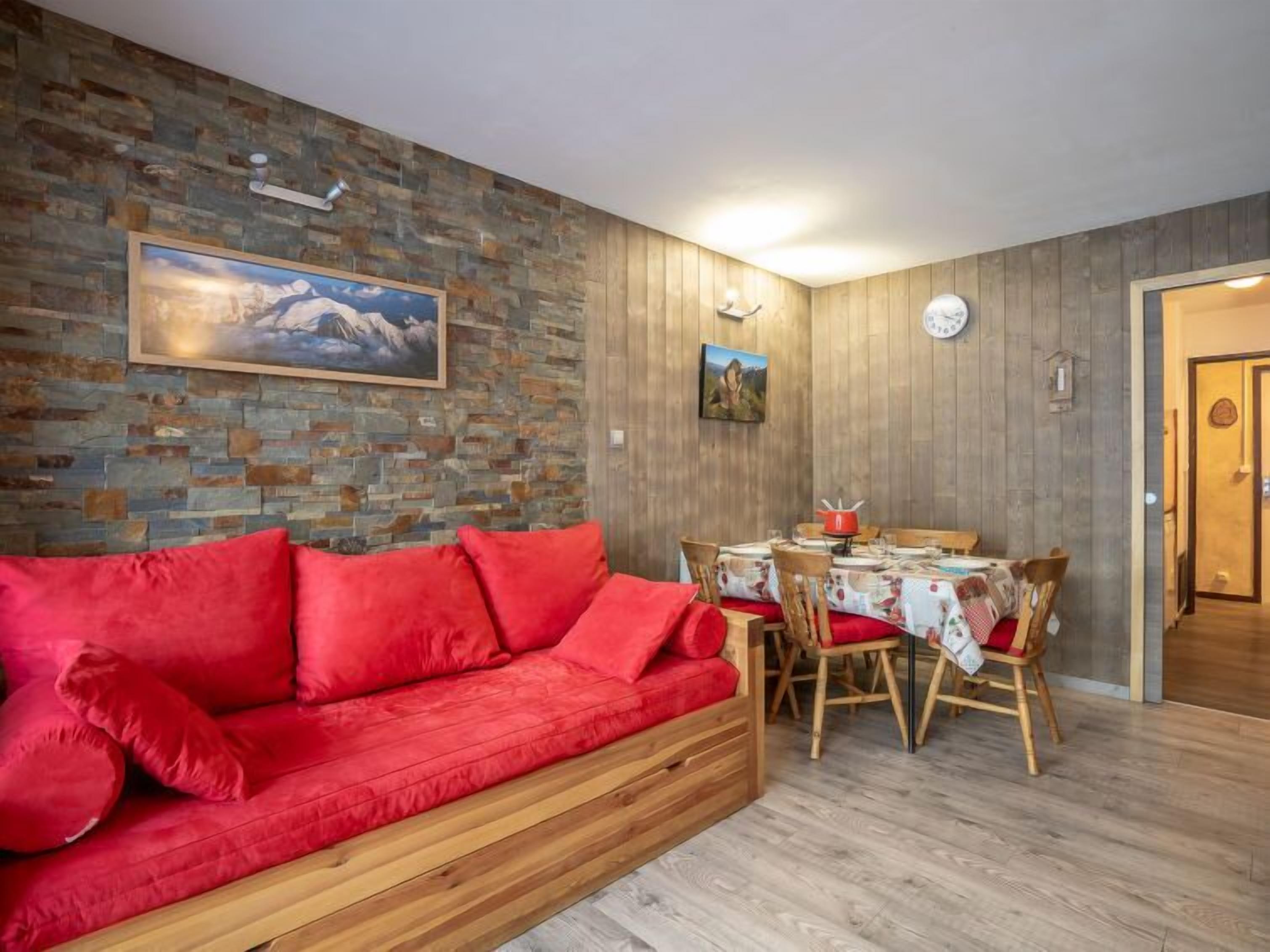 Ein gemütliches Wohnzimmer mit einem roten Sofa, einem Holzespeisetisch und Stühlen. Ein Gemälde hängt an der Wand, und eine Uhr ist sichtbar.