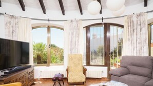 Una sala de estar con televisor, sofá, mesa de centro de vidrio y una silla. Hay ventanas grandes con cortinas, un armario de madera y un radiador.