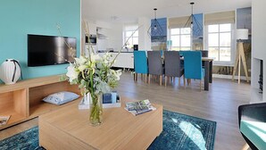 Ein modernes Wohnzimmer mit Flachbildfernseher, Couchtisch mit Blumenvase und Essbereich mit blauen Stühlen und einer Chandelüster.