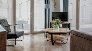 Una sala moderna con un sofá, un sillón, una mesa redonda de madera con un jarrón de flores, un televisor sobre un mueble bajo y ventanas amplias con persianas.